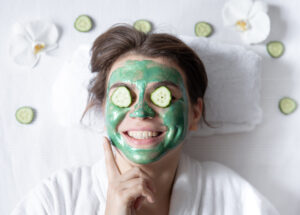 Funny young woman with a cosmetic mask on her face and cucumbers on her eyes.
